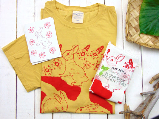 A yellow unisex t-shirt with a red rabbit print, accompanied by a matching red and white dish towel with a rabbit design, and a note card. The t-shirt and tea towel are displayed flat with a woven basket in the background.